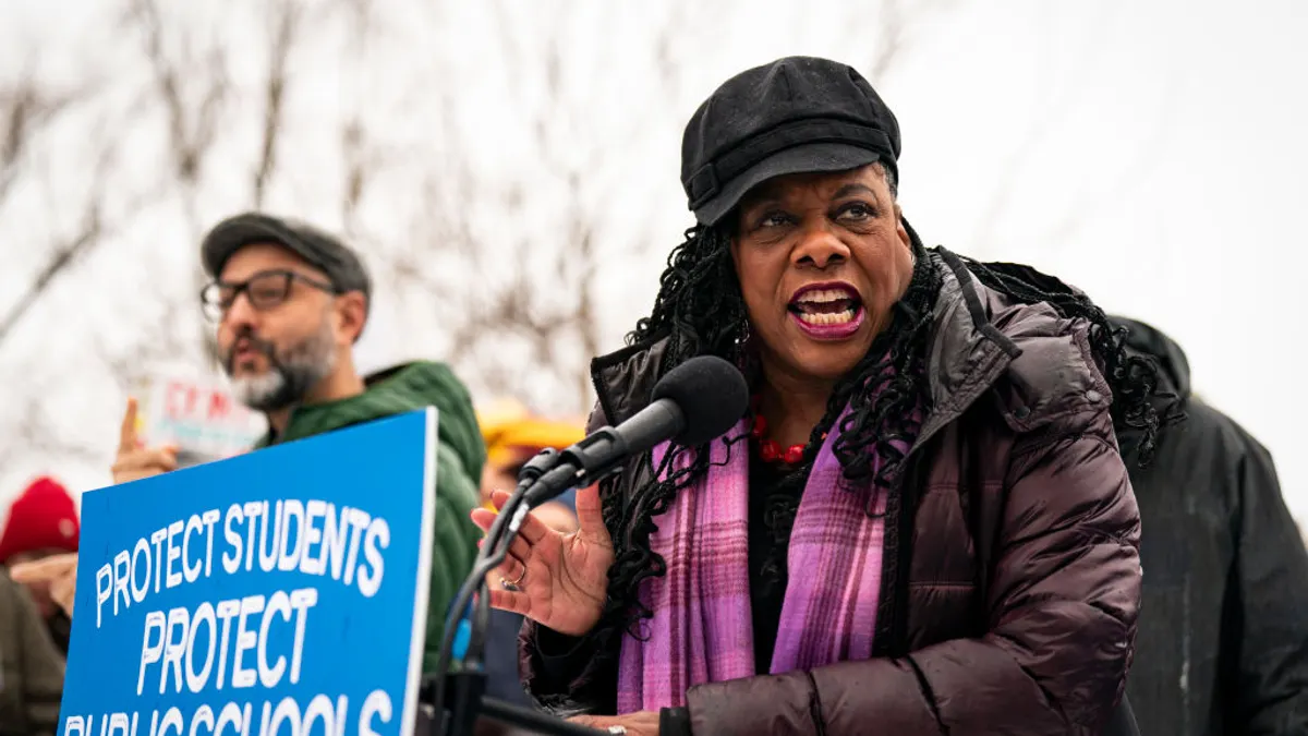 A person wearing a black hat and purple jacket and scar speaks into a podium microphone on a winter day