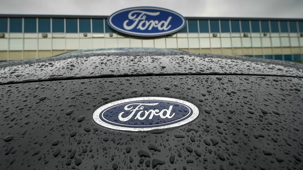 Ford Motor logo is seen on the hood of a blue car. In the background, a manufacturing plant with the Ford logo can also be seen.