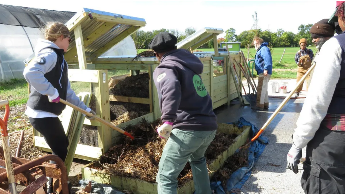 Two people wielding shovels move organic material from a pallet to a composting bin while others look on.