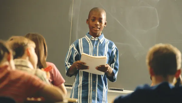 A student is holding a notebook and is standing in front of a classroom. Classmates are sitting at desks facing the student.