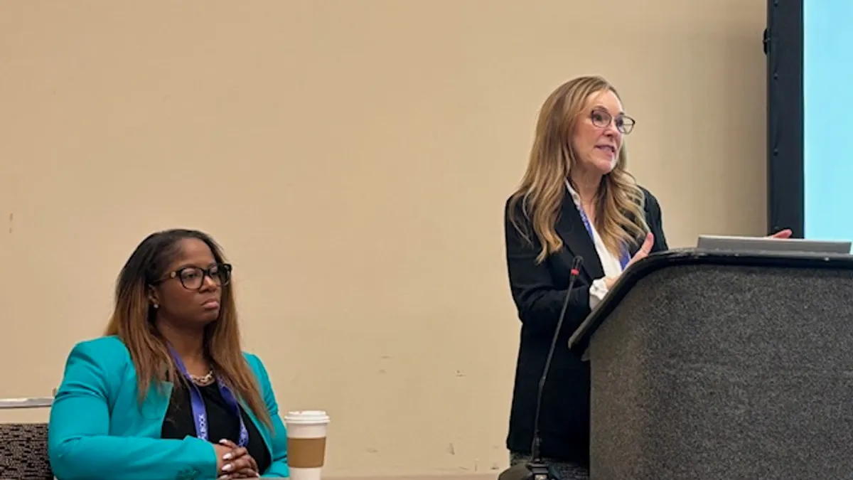 Two people are pictured. One is standing behind a podium in a room and the other is sitting at a desk next to the podium