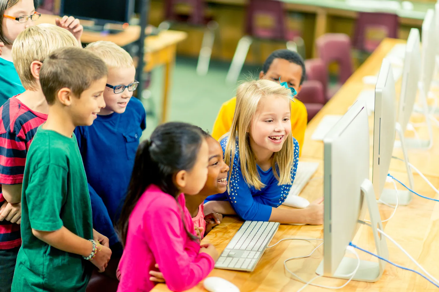 A handful of young students are in a room with desks. They are looking at one computer monitor on the desk that also has other computer monitors