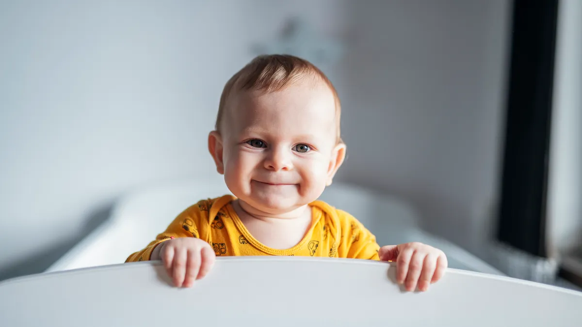 A baby stands in a crib, smiling and looking at the camera.