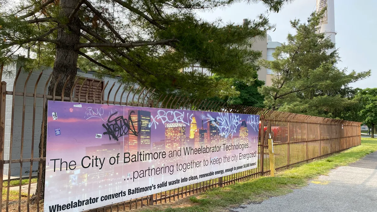 A lightly graffitied sign outside an industrial facility reads "The City of Baltimore and Wheelabrator Technologies ... partnering together to keep the city Energized!"