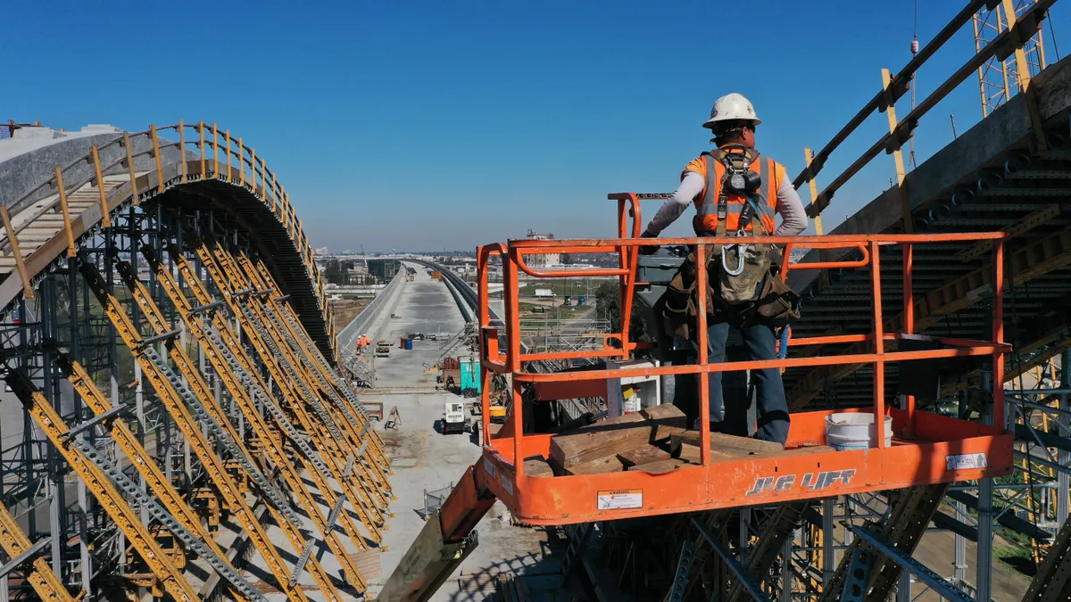 A construction worker stands in a cherry picker high above a worksite, where a long concrete viaduct is visible.