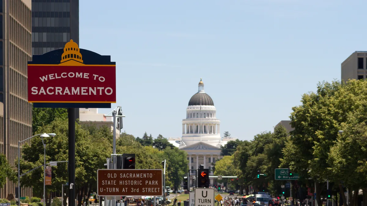 A sign that welcomes visitors to the city of Sacramento sits in the foreground, in front of a tall, white, domed building. Ahead is a long shot of grass, with buildings on the periphery.