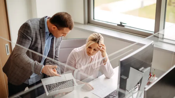 Two colleagues have a discussion at an office desk as one points to a laptop screen.