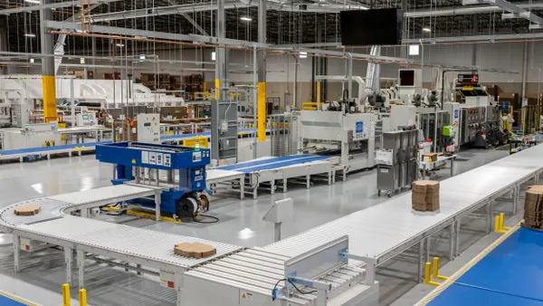 A view inside a manufacturing facility shows heavy equipment and corrugated boxes on conveyors.