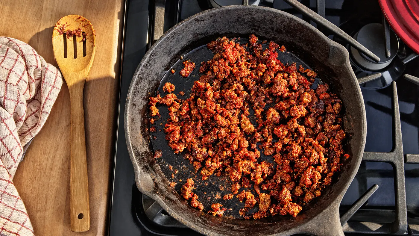 A cast iron skillet with cooked Impossible Beef Lite sits on a range with a wooden slotted spoon that was used to saute it sitting to the left.