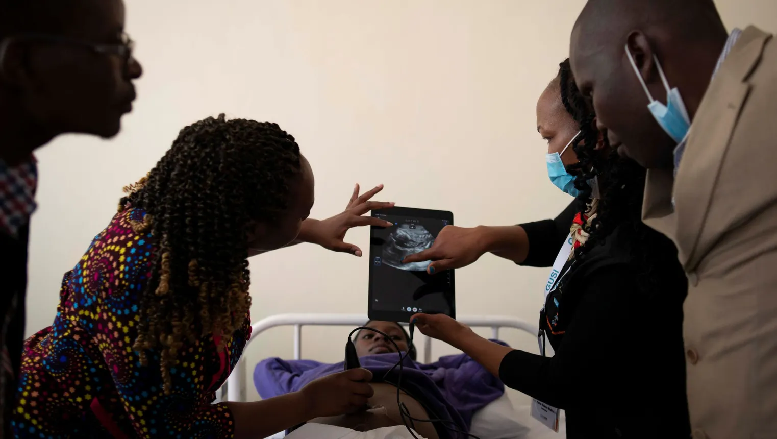One person is screened by an ultrasound, while four people look at a tablet depicting the image.