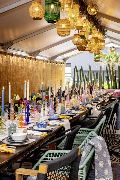 A row of chairs seated at a colorful, long table with candlesticks on it and lamps hanging down from above.