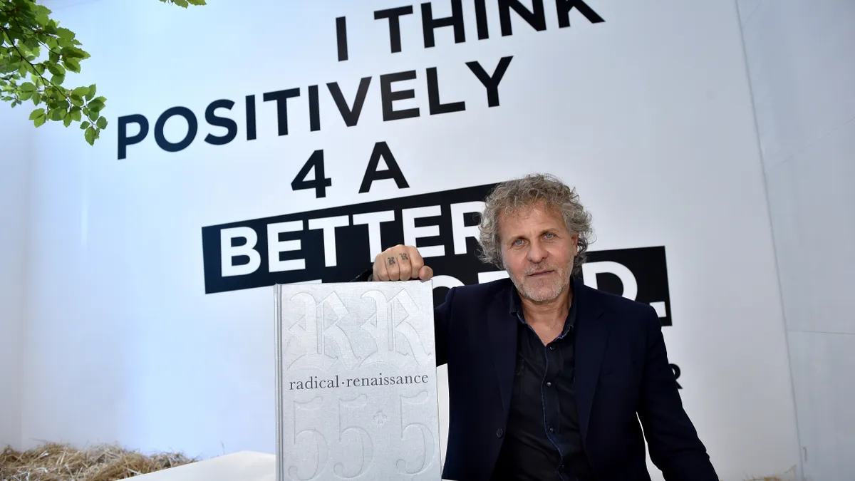 Man with grey hair wears a white suit and sits at a white table holding a large white book in front of black and white lettering reading "I think positively for a better world."