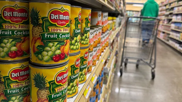 Cans of Del Monte fruit cocktail on a shelf in a grocery store with a shopping cart and customer nearby.