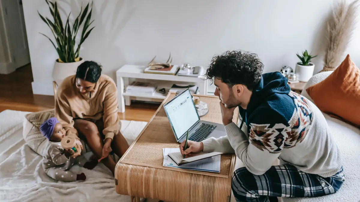 A working parent looks intently at their laptop, while their partner plays with their baby