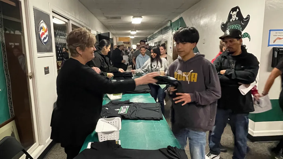 People are standing in a school hallway. A table is in the hallway and on the table are T-shirts with the word Pirates. A person is handing another person a T-shirt.