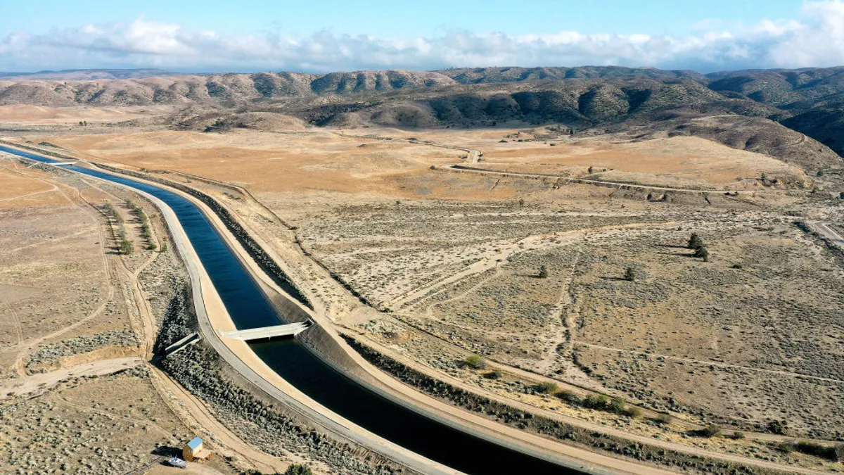 Aerial view of the California Aqueduct, a ribbon of water running through the desert with mountains in the background.