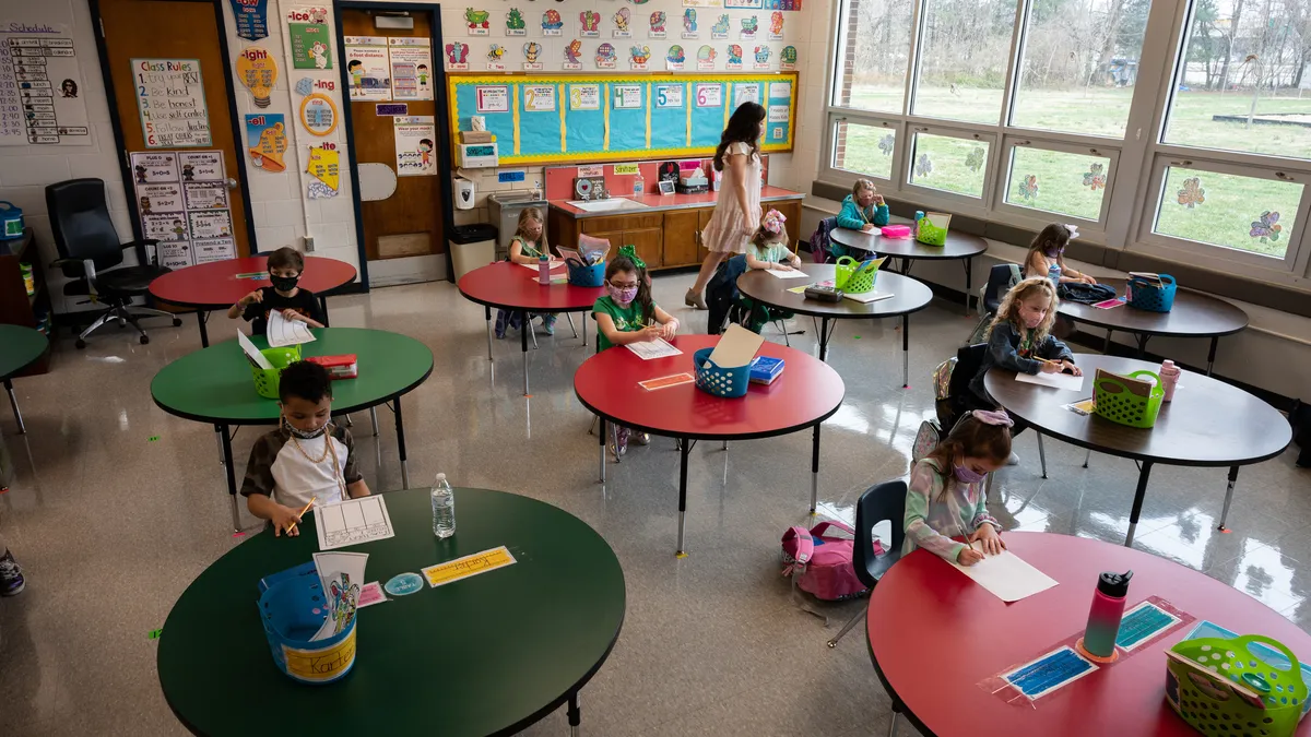 Students sit at tables in classroom with teacher walking.