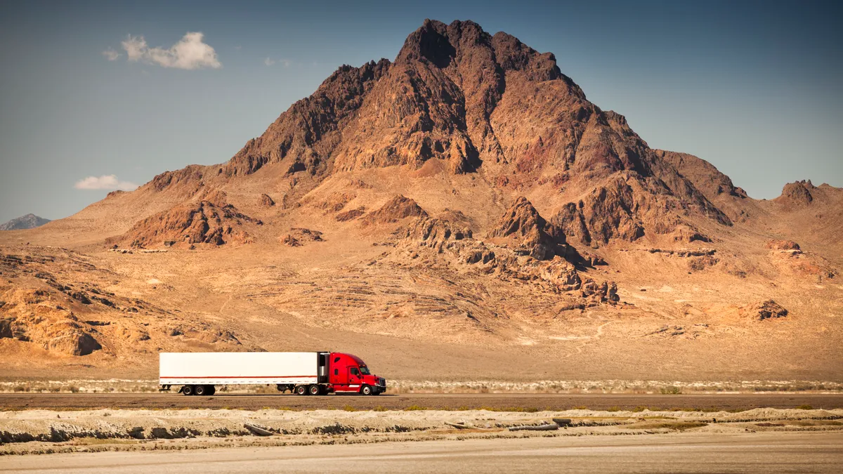 A tractor-trailer moves along a road by a barren, rocky mountain.