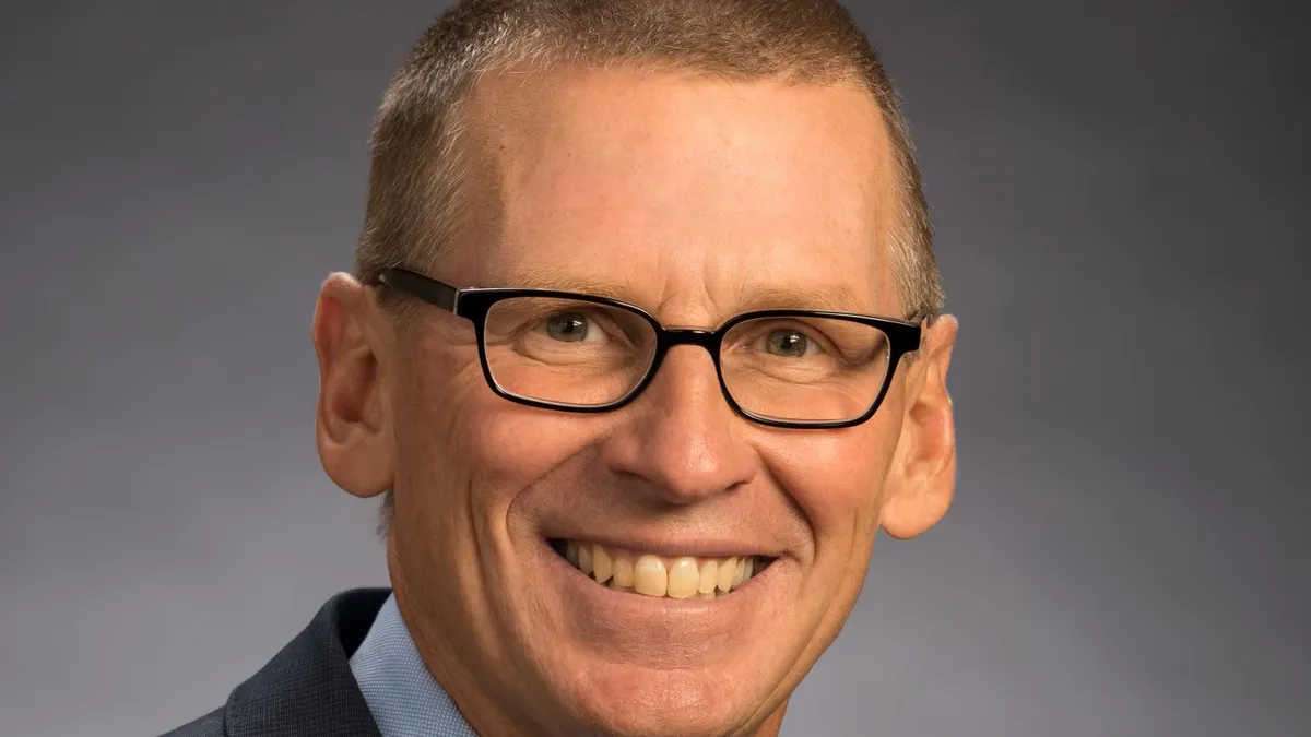 A photograph of a smiling man wearing a black suit, light blue shirt and blue tie.