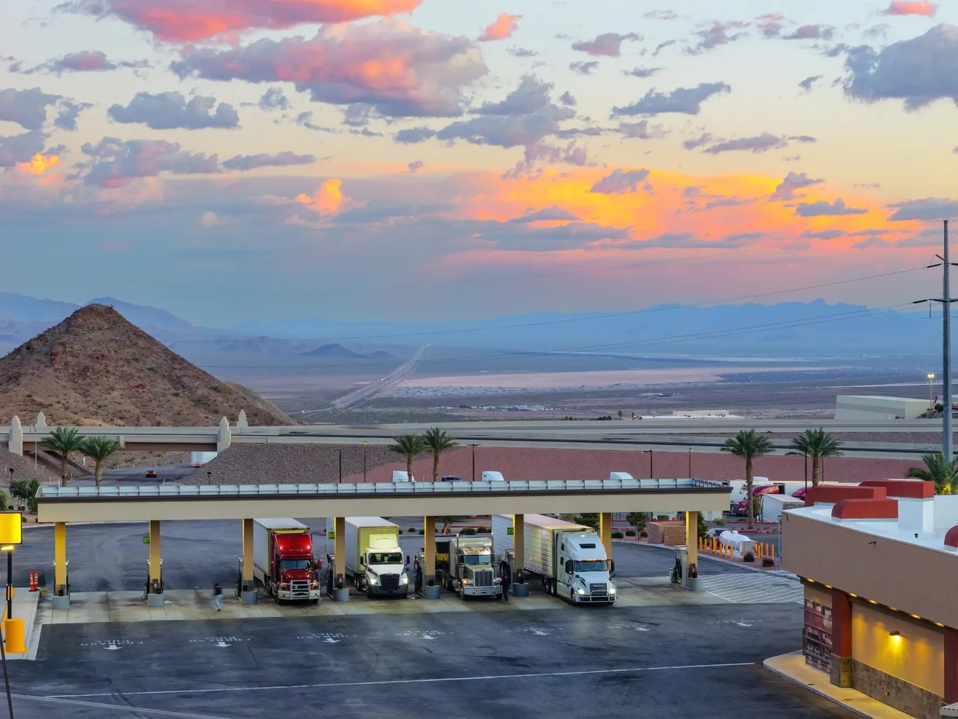 Trucks fueling against a dramatic evening sky showing sunlight across clouds amid a desert landscape.