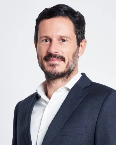 A person with short dark hair, beard and mustache smiles, wearing a white shirt and jacket but no tie.