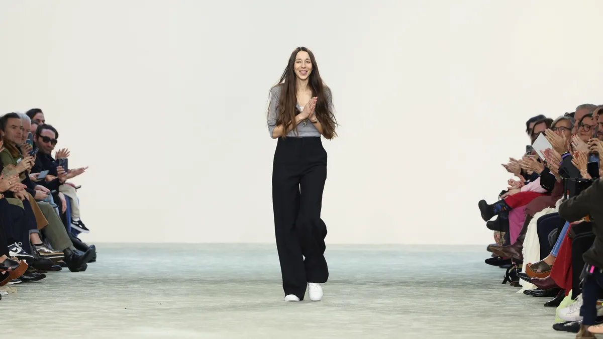A person with very long, straight hair stands at the end of an empty runway with clasped hands.