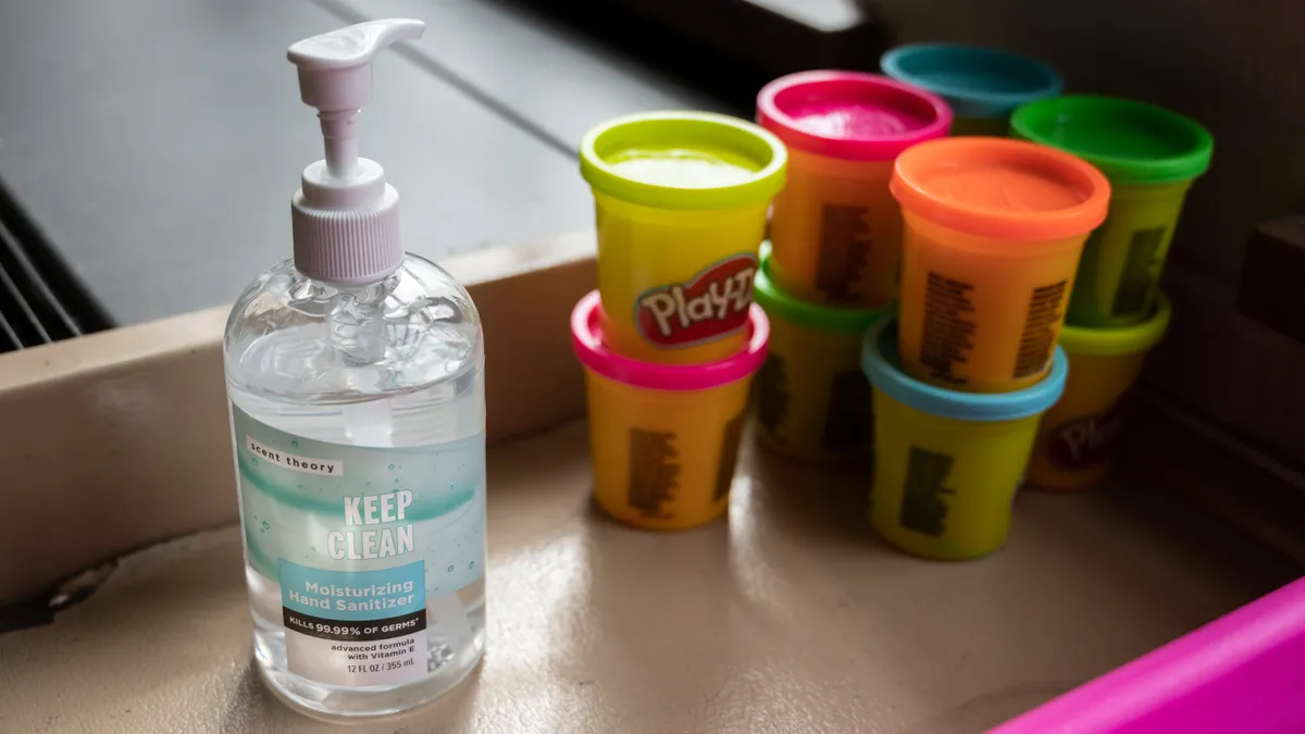 A pump bottle of hand sanitizer sits on a desk next to stacked cans of colorful Play-Doh.