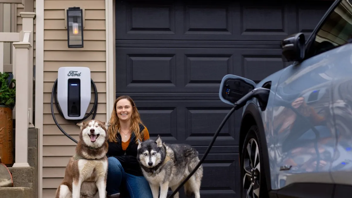 A person kneels with two large dogs in front of a garage with a Ford residential EV charger mounted on a wall.