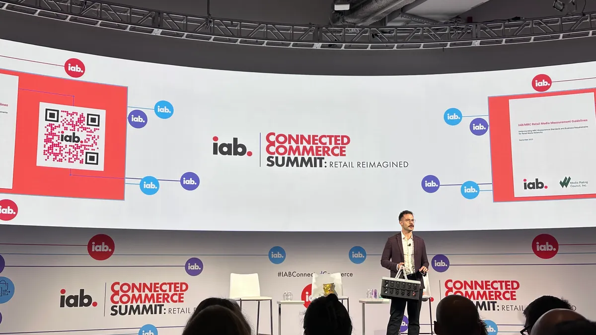 Man on stage with gray grocery basket giving a keynote speak at IAB's Connected Commerce Summit, which is written on a projector screen above