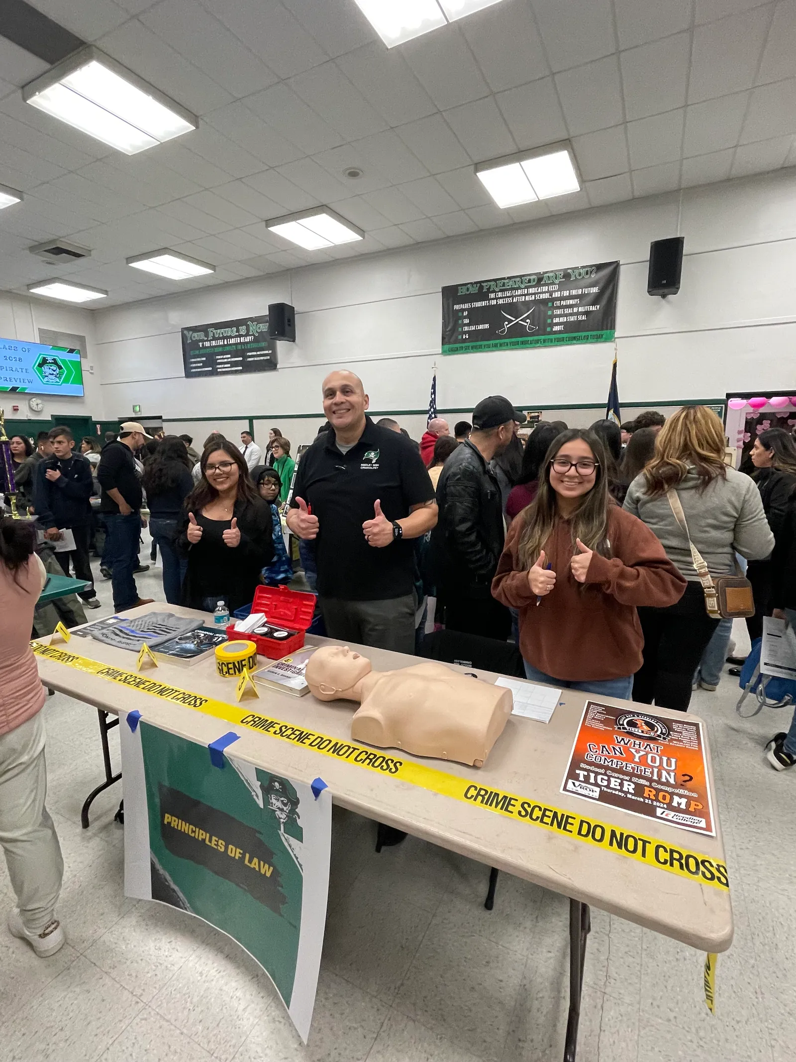 People are standing behind a table in a school gymnasium. They are looking at the camera and holding their hands up with the thumbs up sign.