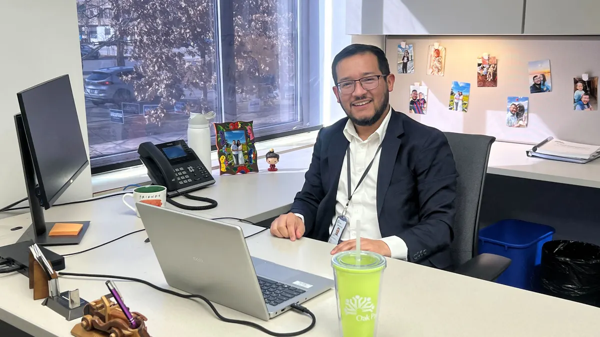 A person dressed in business attire sits at a office desk with a laptop.