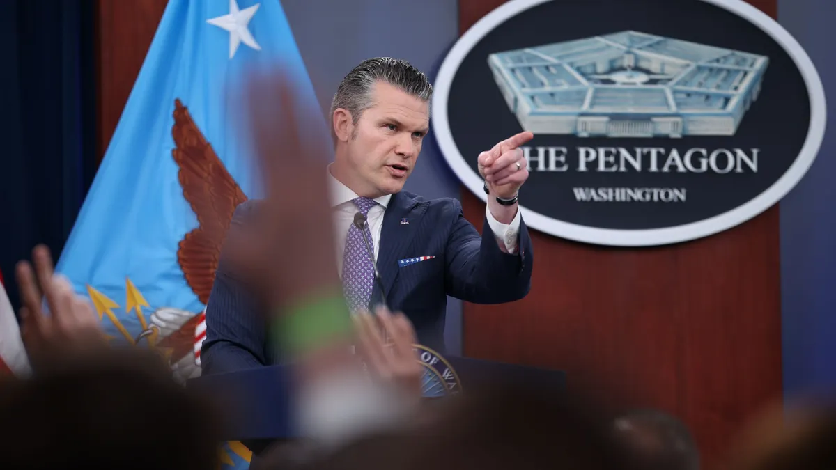 A man in a suit standing behind a podium pointing, with the oval blue and white Pentagon Washington sign in the background.