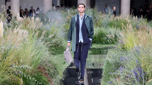 A models walks on a runway wearing a casual suit. On the sides of the runway are tall plants and weeds.