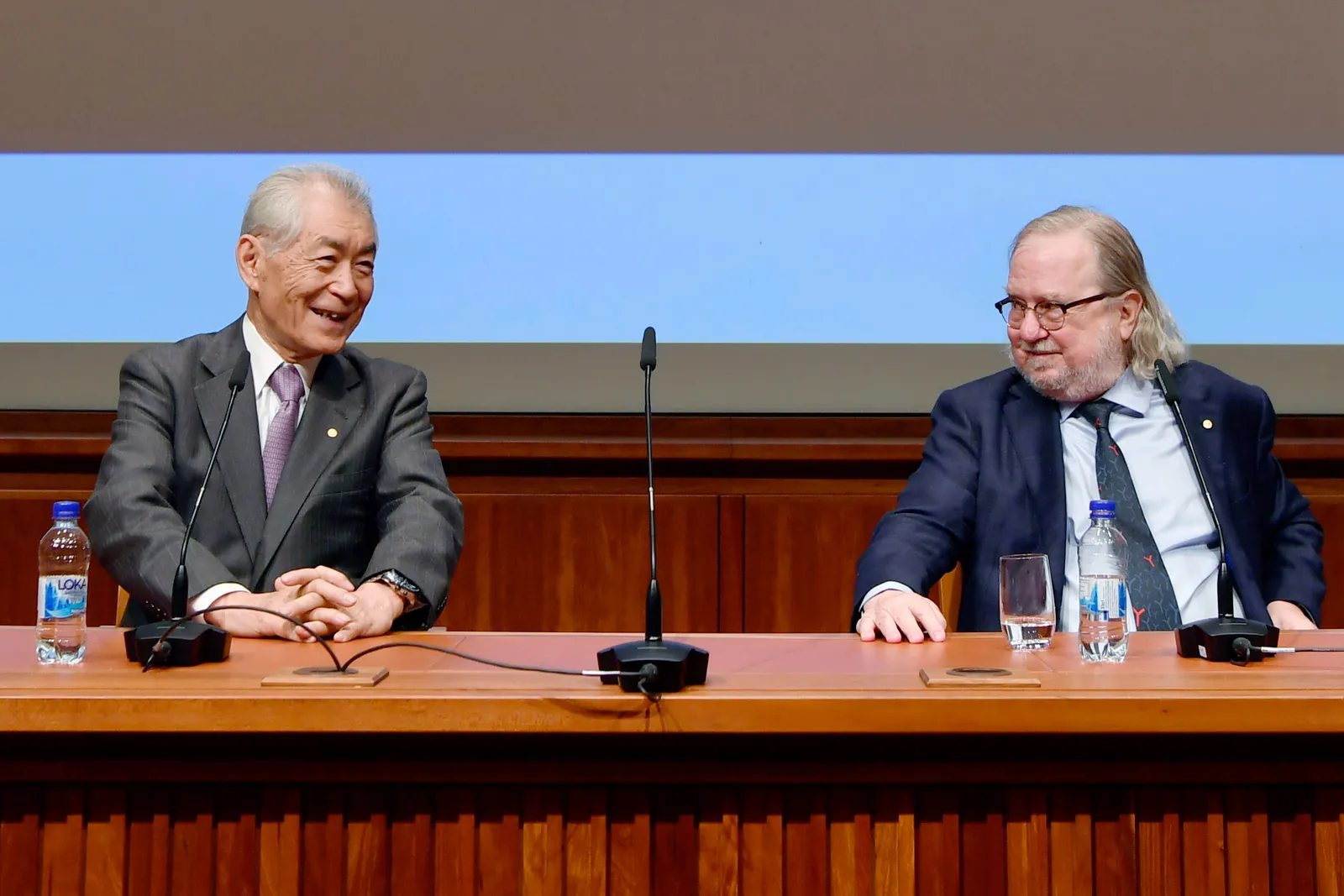 Two people in business attire sit behind a wooden dias with microphones in front of them.