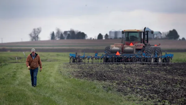 A farmer in a baseball cap walks alongside a plowed field. In the distance, a tractor is seen.