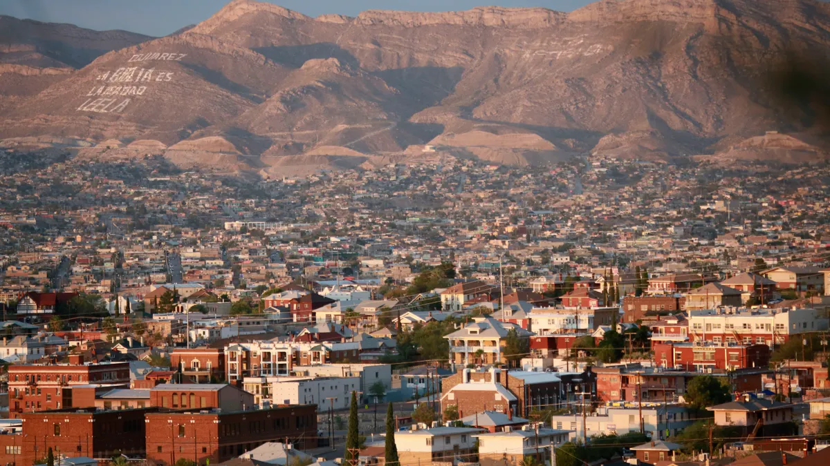 An aerial view over El Paso, Texas shows tightly-packed buildings in the foreground with brown hills in the background.