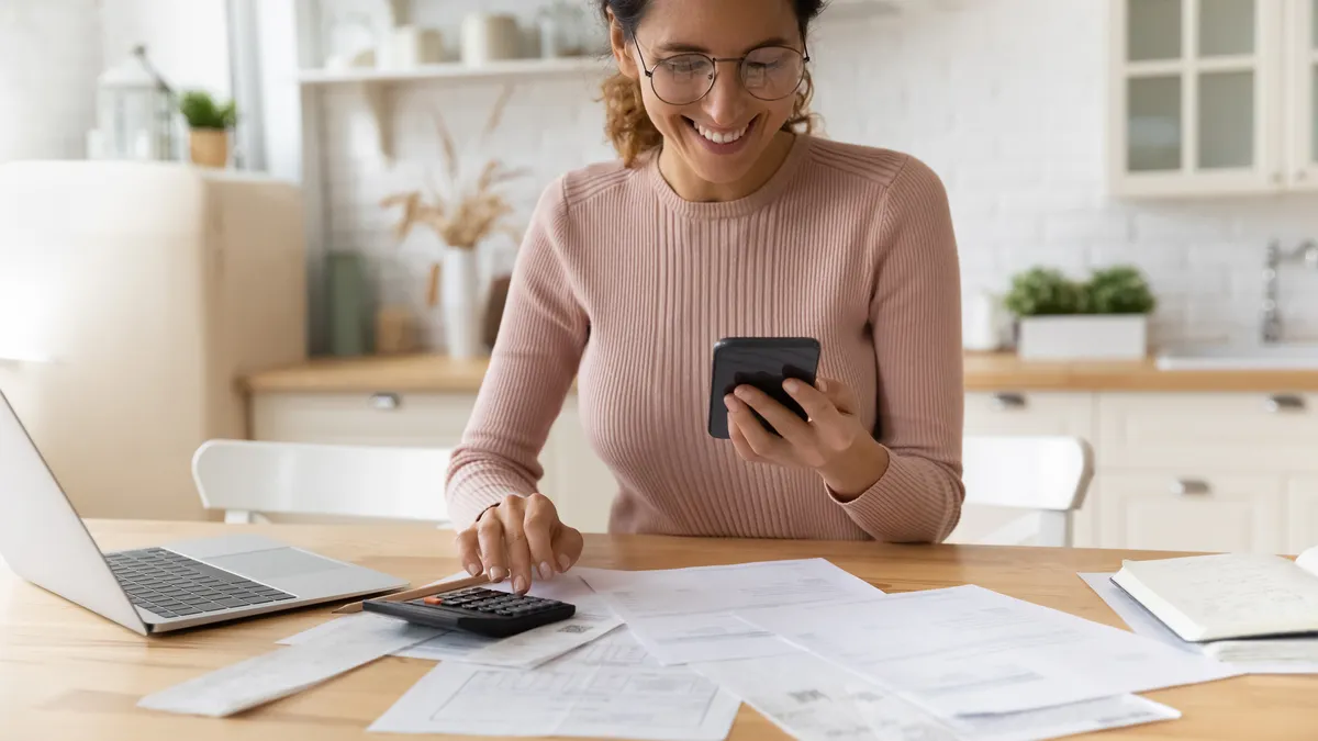 A woman sits at a desk, looking at her phone and doing paperwork, seemingly to pay bills.