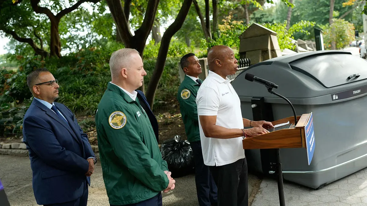 Eric Adams stands at a podium in a public park on a sunny day. Behind him are several officials and a large bin, roughly five feet tall with a handled lid and a "Trash" label.