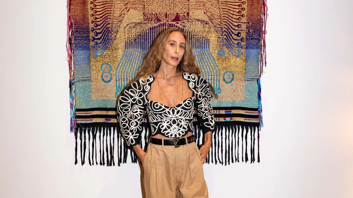 A person stands in front of a tapestry wearing gray pants.