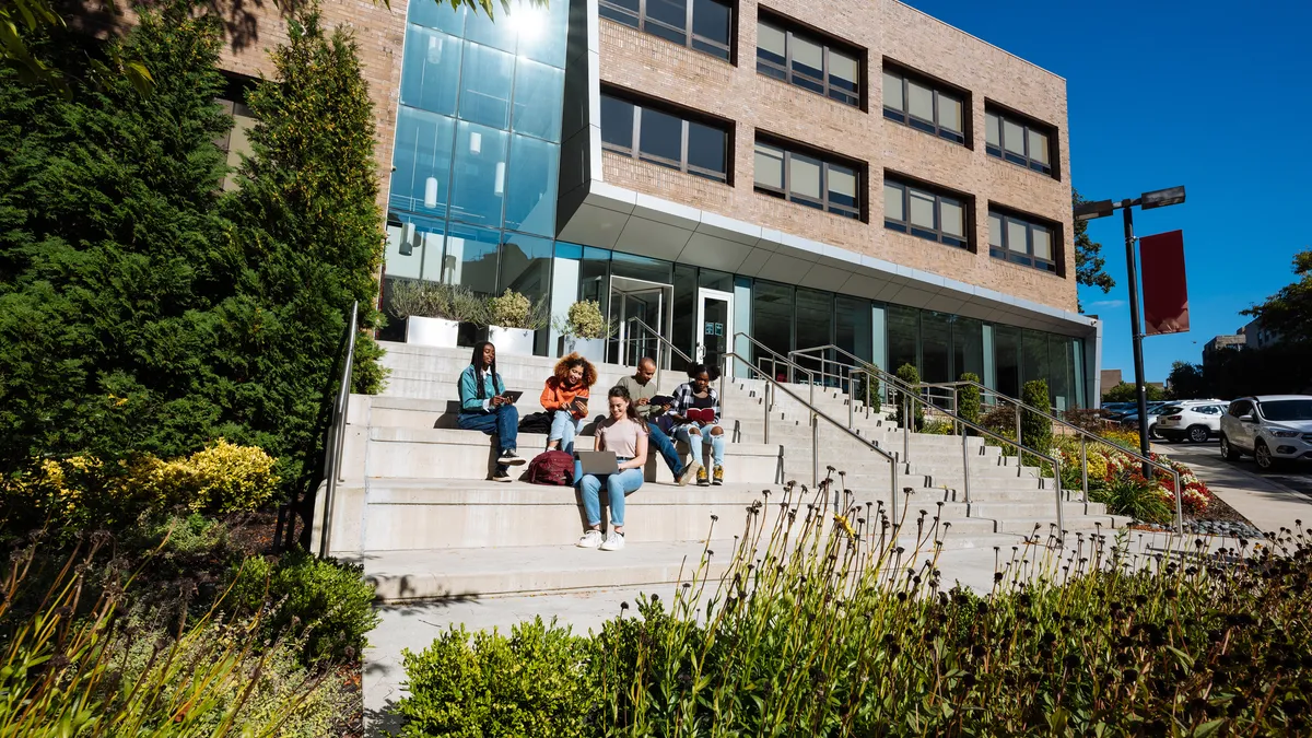 A group of college students sit on a building's steps with their laptops open.