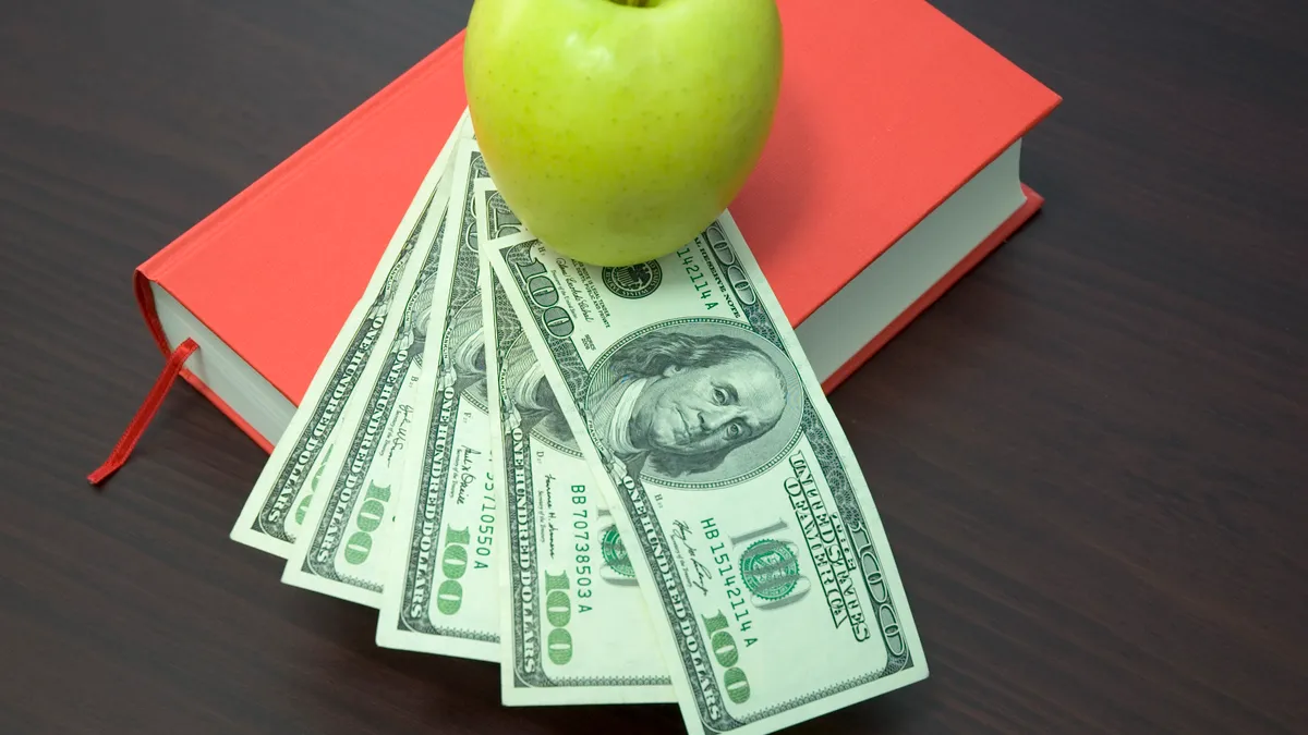 A green apple sits on top of a fan spread of $100 bills, which sits on top of a red book.