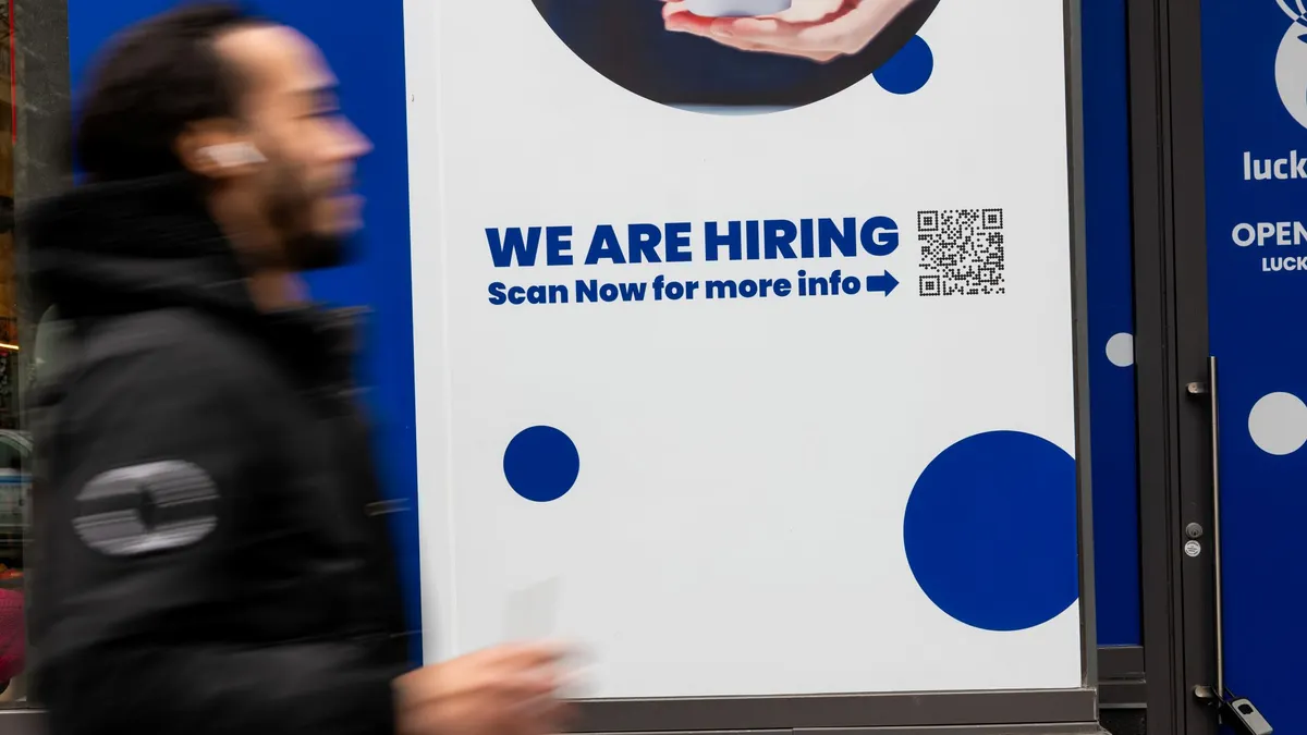 A person walks by a "now hiring" sign bearing a QR code.