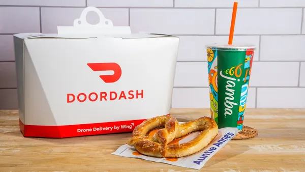A white container with a DoorDash logo sits on a table next to a green cup with a Jamba logo and a pretzel.