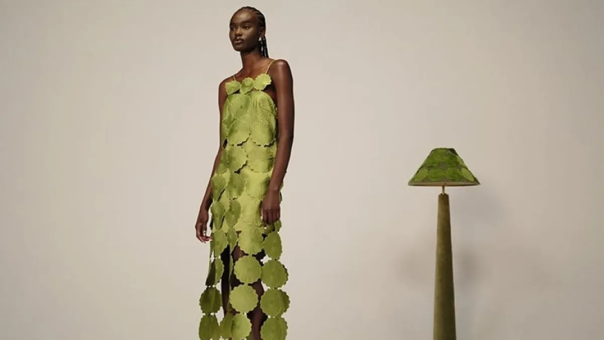 A person in a dress made of green circles stands next to a green floor lamp.