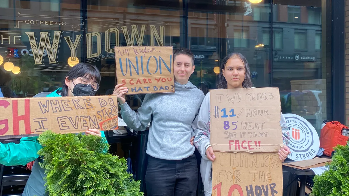 Three people stand outside a closed coffee shop holding signs.