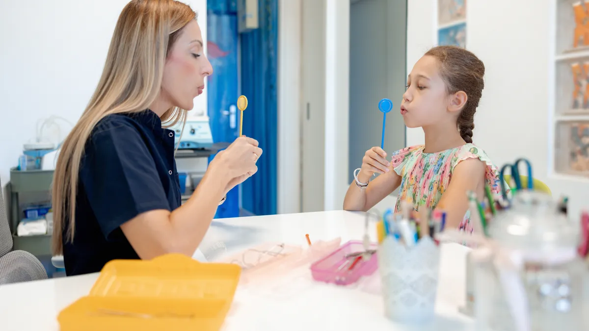 An adult speech therapist works one-to-one with a student at a desk. Both are holding a tool in front of their faces and blowing on the tool.