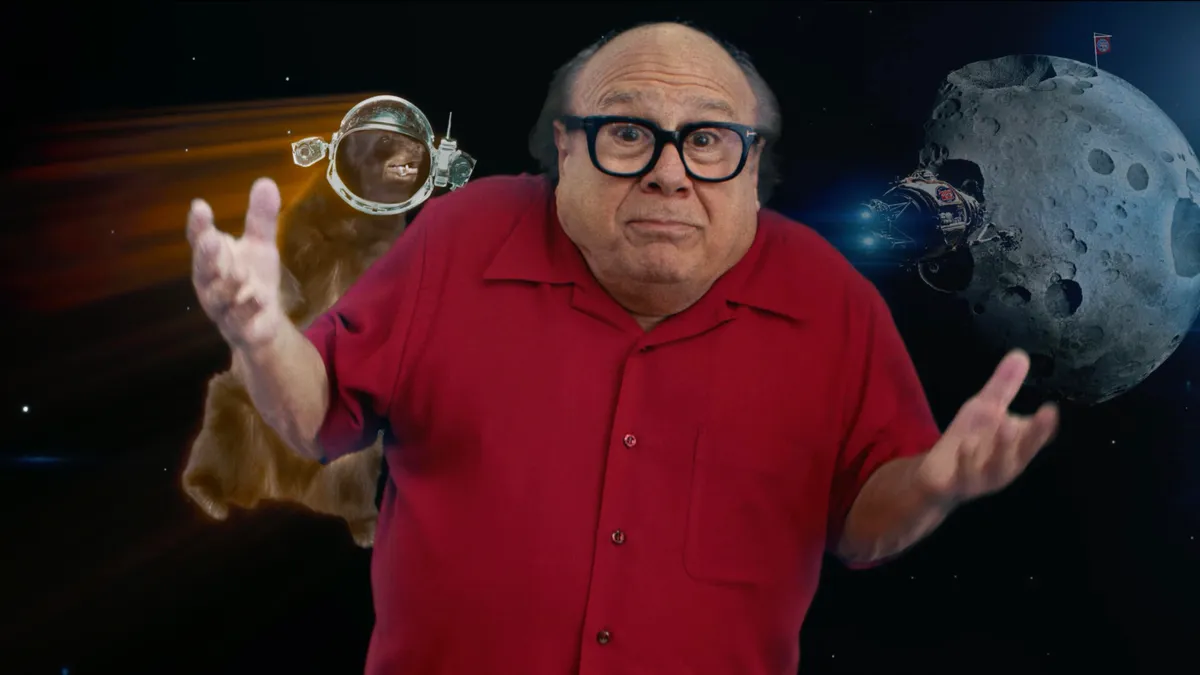 Danny DeVito stands in front of a green screen image of space.