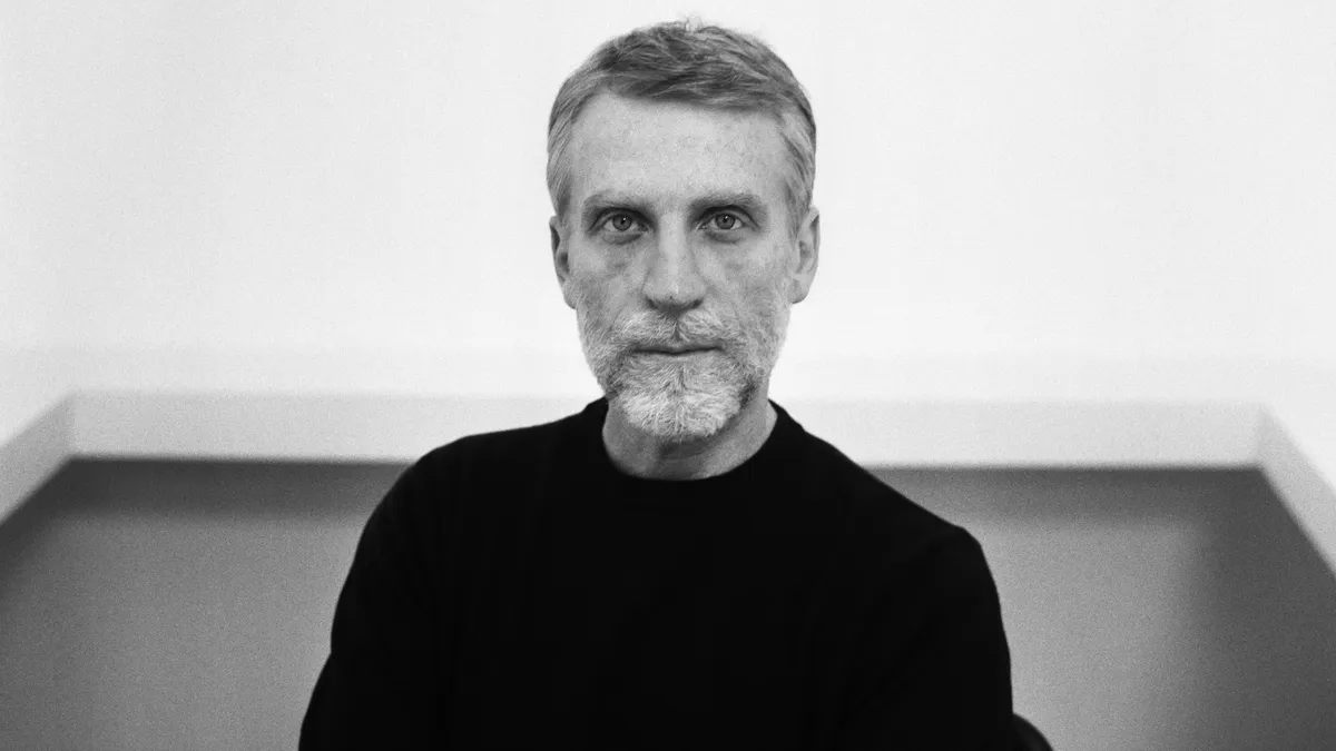 In a black-and-white corporate headshot, a person with a bread and mustache wears a black turtleneck and stares at the camera.