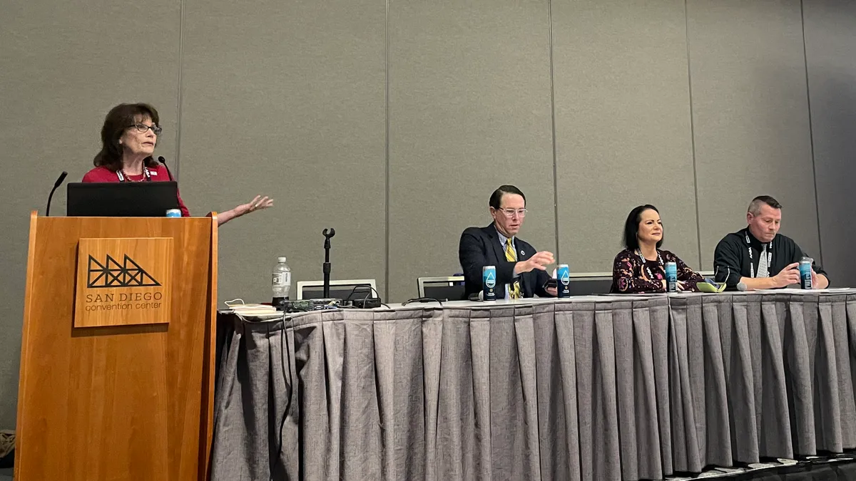 A group of three school superintendents are seated at a table on a dais in a convention center meeting hall for a panel session. A presenter stands at a podium to the left.