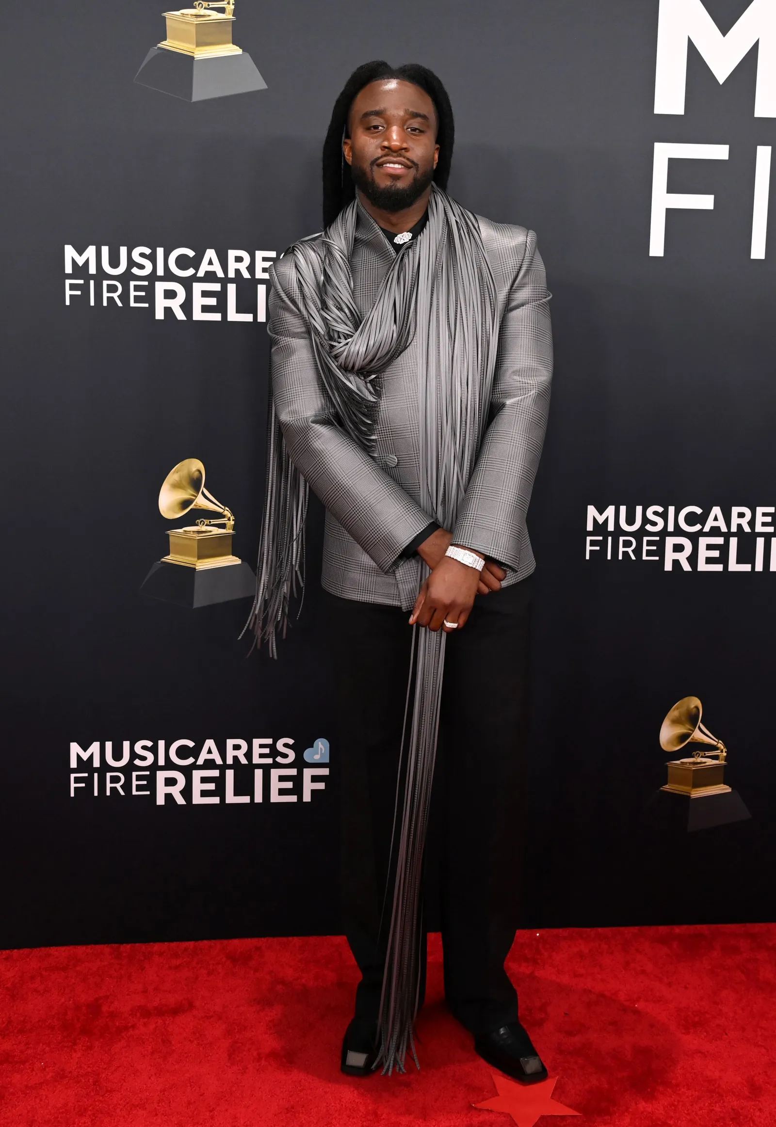 A person in a silver jacket and fringe scarf stands on a red carpet.